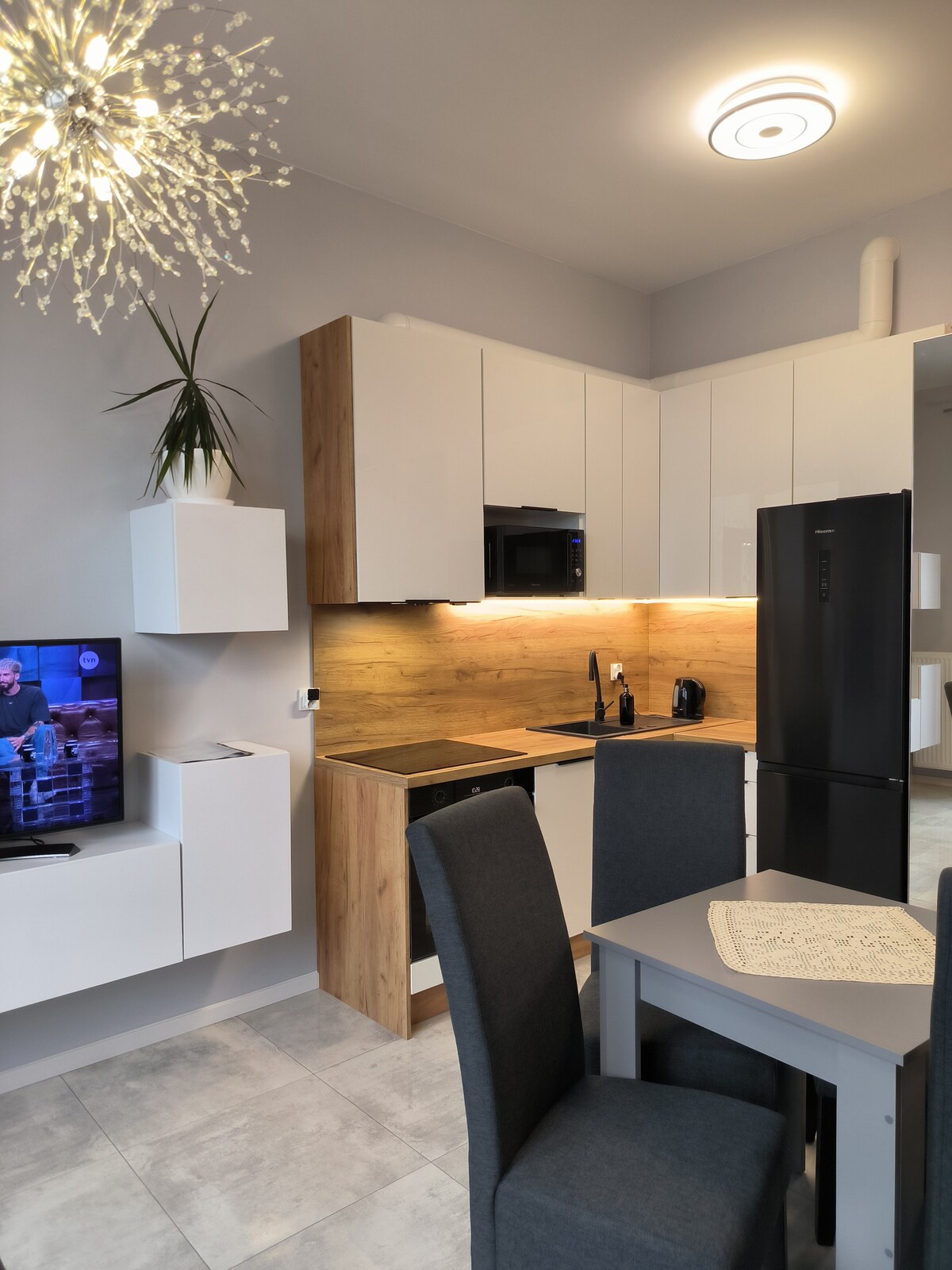 A modern kitchen area is equipped with essential appliances, including a refrigerator and microwave. Light wood and white cabinetry creates a clean aesthetic. A dining table with gray chairs is positioned nearby, with a decorative light fixture enhancing the ambiance of the space.