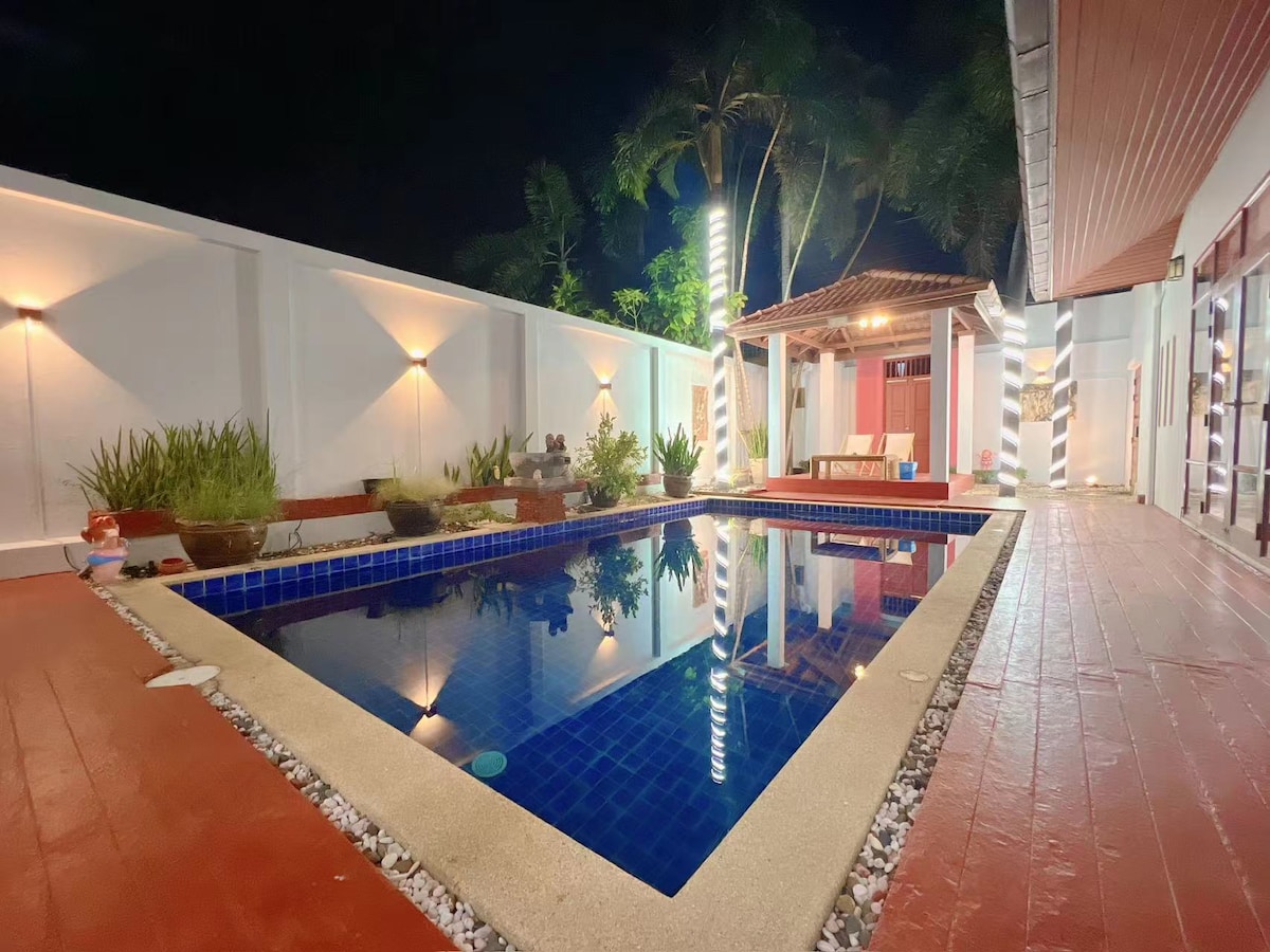 A private swimming pool is illuminated by soft lighting at night, reflecting the surrounding tropical greenery. A gazebo with seating is positioned nearby, creating a serene outdoor space for relaxation. Red and white accents on the patio enhance the inviting atmosphere.