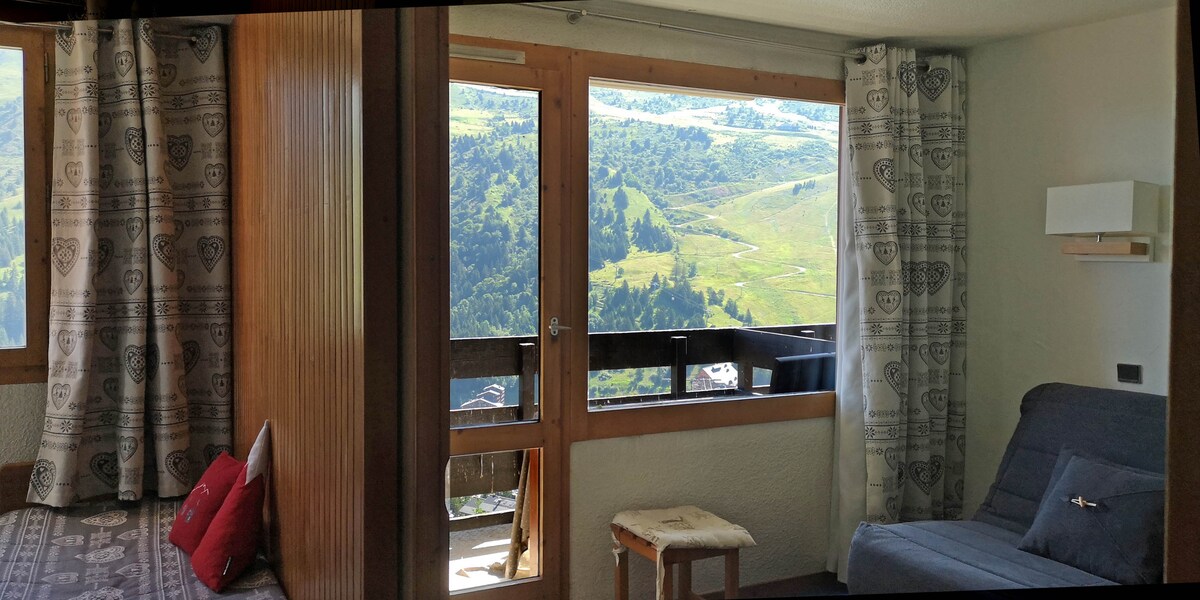 A cozy living area features large windows that offer expansive views of the mountains and valleys. A simple sofa bed is positioned next to a light wooden side table. Curtains with a subtle pattern frame the windows, enhancing the space's natural light.