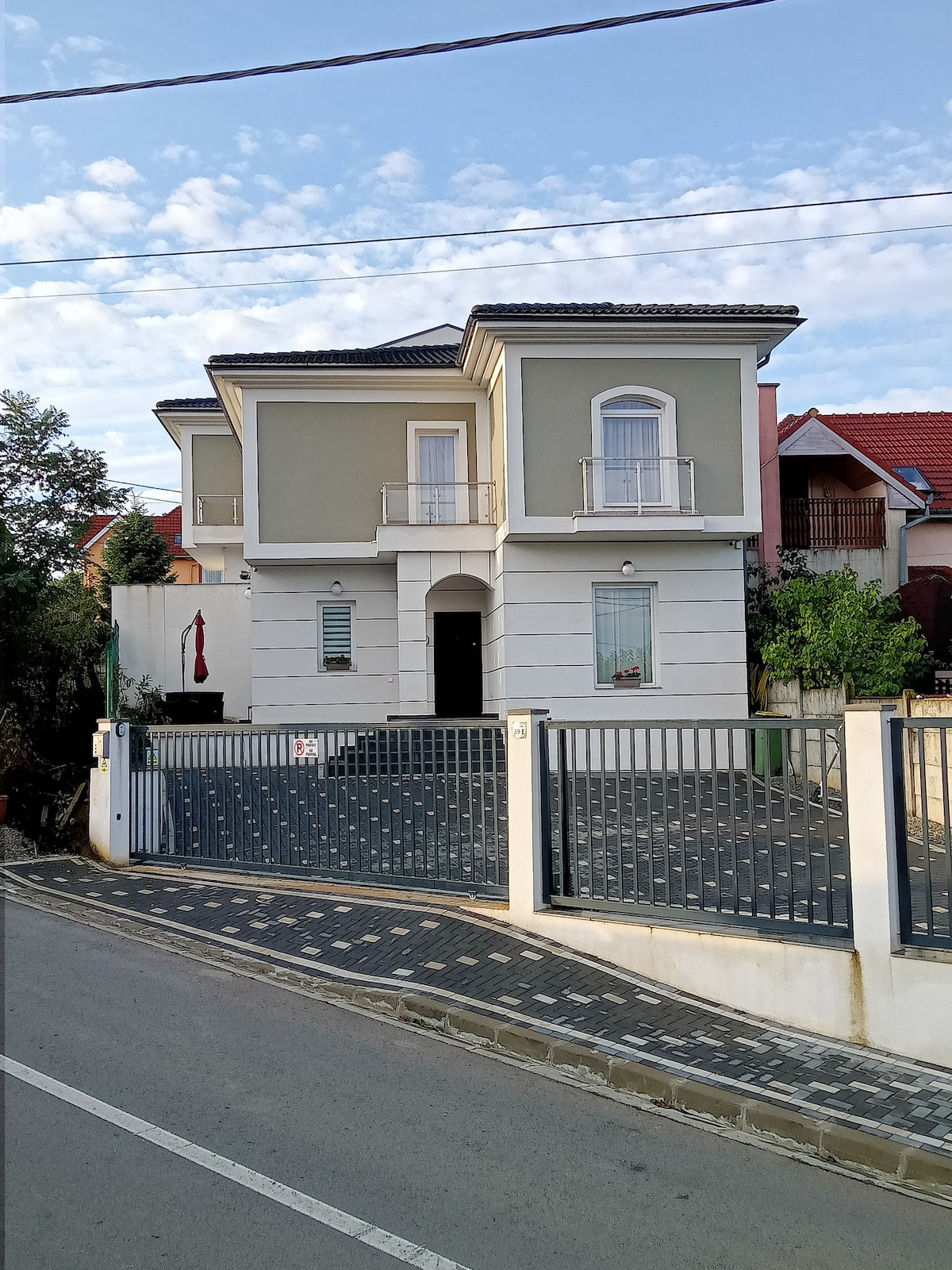 A two-story house is presented with a light green and white facade. The entrance is framed by symmetrical windows and a balcony above. A gated driveway leads to a spacious parking area, with landscaped greenery on either side.