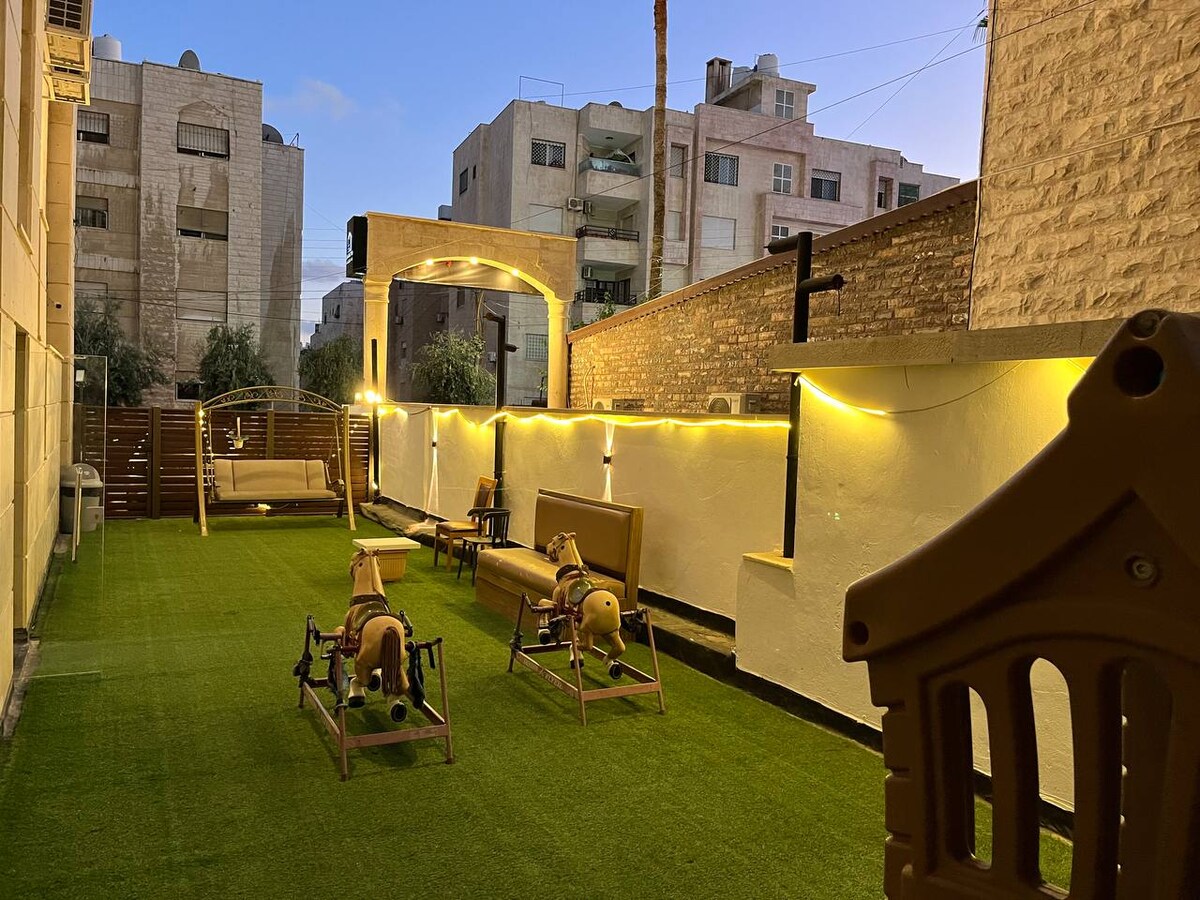 An outdoor area features artificial grass with two wooden rocking horses positioned on either side. Wooden benches and swings are situated against the wall, illuminated by ambient lighting. The space provides a quiet environment, bordered by neighboring buildings.