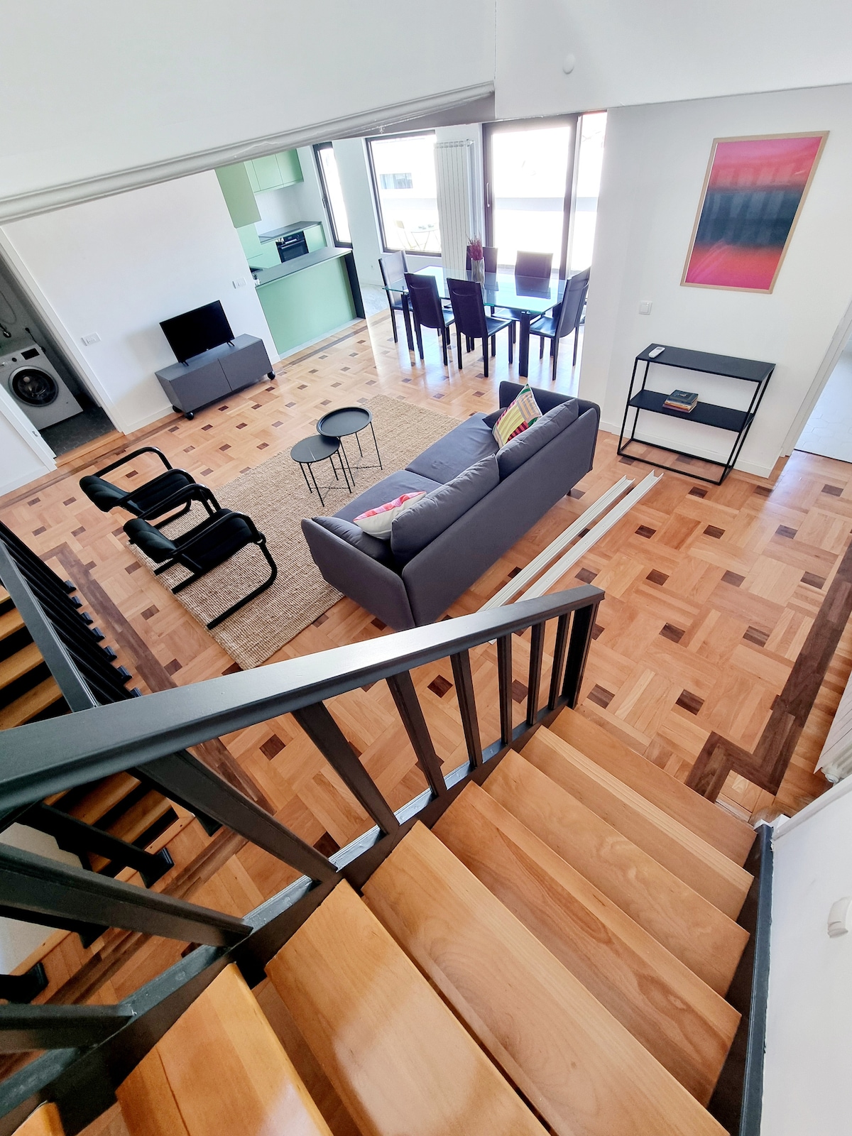 An open staircase leads to a spacious living area featuring wooden flooring. A comfortable gray sofa and two chairs are arranged around a small coffee table. Nearby, a dining table is visible, accented by large windows allowing natural light to fill the space.