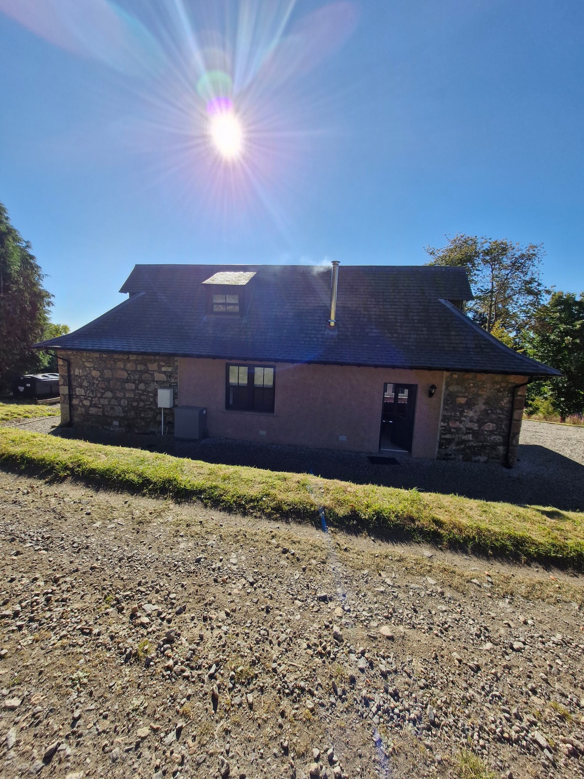 A charming cottage with a mix of stone and smooth surfaces is set against a clear blue sky. Sunlight creates a bright reflection on the roof, while the front entrance is visible. The surrounding grounds feature gravel paths and greenery, contributing to a peaceful ambiance.