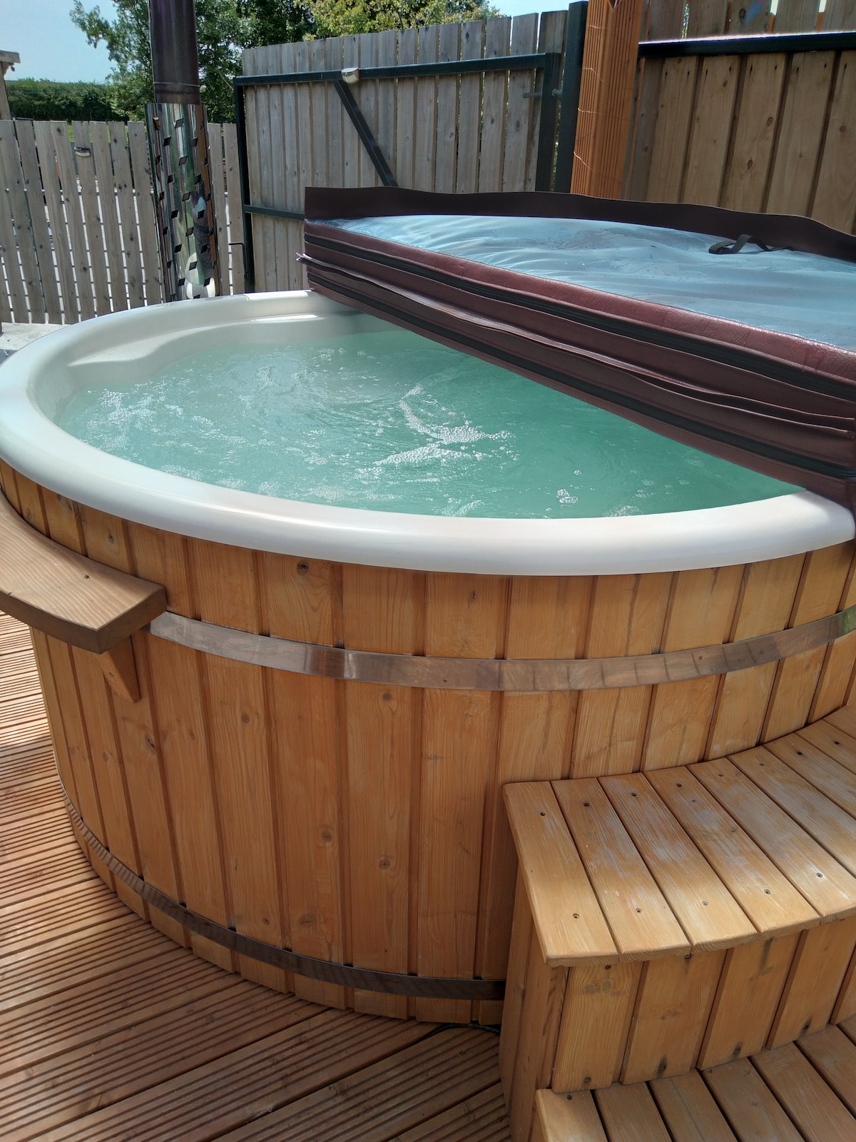 A wooden hot tub is shown, featuring a smooth, rounded edge and clear water. The tub is accompanied by steps for easy access. Surrounding elements include wooden decking and a backdrop of wooden fencing, offering a sense of privacy.