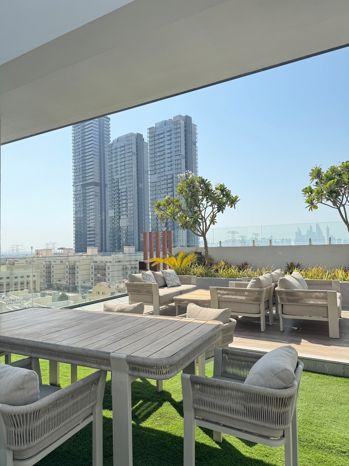 A spacious balcony is showcased with a modern outdoor dining table and four chairs. Lush greenery and stylish seating areas are visible, complemented by a view of tall buildings in the background. The clear blue sky enhances the open atmosphere of the space.