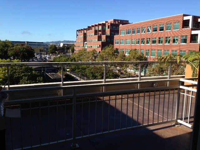 An outdoor balcony is displayed, featuring a metal rail that offers a view of nearby brick buildings and green trees. The skyline is partially visible, with a clear blue sky overhead, providing a sense of openness and connection to the urban environment.
