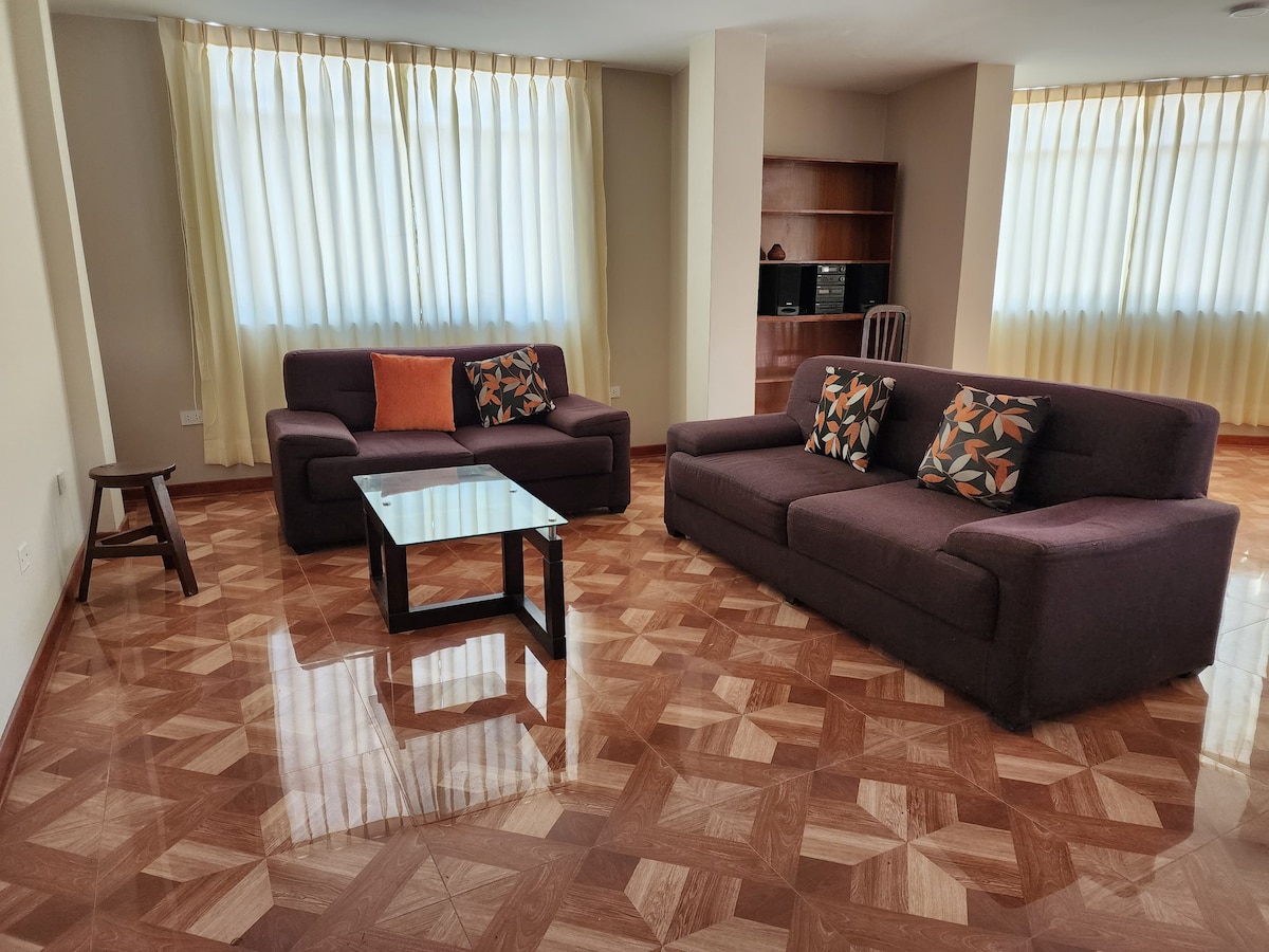 A spacious living room is presented with two comfortable sofas adorned with colorful cushions. A coffee table is positioned in front of the seating area. Natural light filters through large, sheer-curtained windows, enhancing the warm tones of the polished floor.