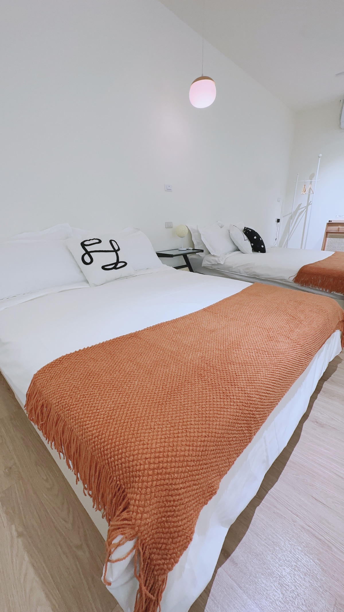 A spacious room features two standard double beds, each adorned with soft white linens and an orange knitted throw blanket. A minimalistic bedside table accompanies one bed, while warm light illuminates the area from a hanging pendant light above.