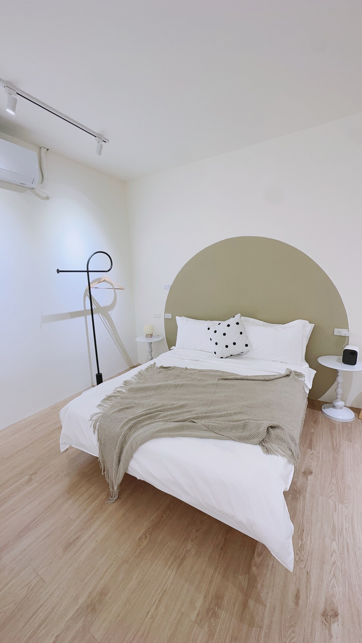 A spacious bedroom features a double bed adorned with a soft blanket and a decorative pillow. A minimalist lamp and small side table complement the serene decor. Natural light enhances the warm wooden flooring and soft wall color.