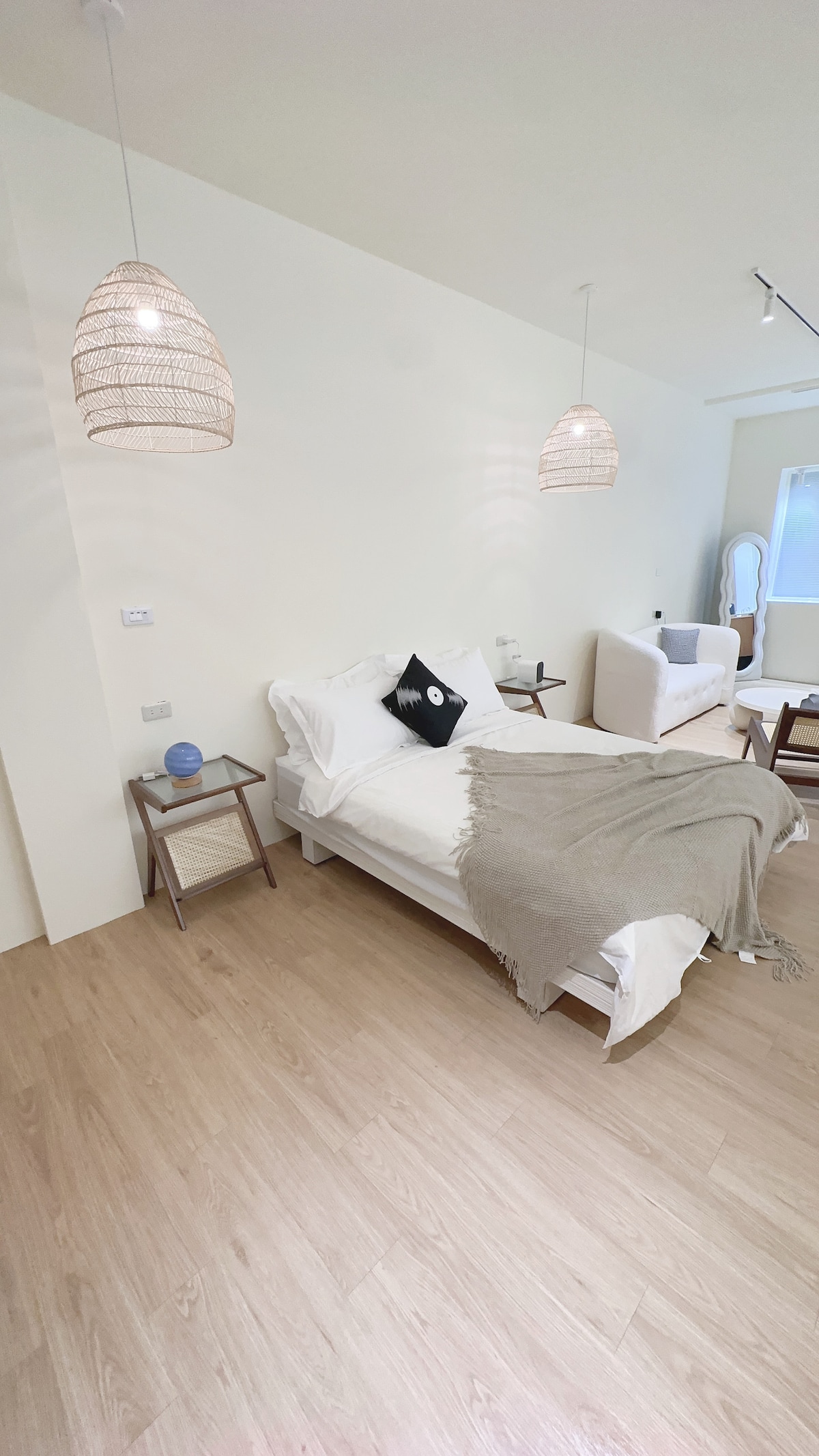 A light and airy bedroom features a spacious bed dressed in white linens, complemented by a neutral-colored throw. Two pendant lights illuminate the space, which includes a small bedside table with a decorative item. A cozy seating area is visible in the background.