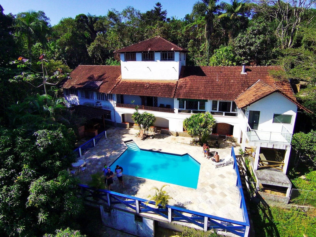 A spacious house is nestled among lush greenery, showcasing a swimming pool that contrasts with the surrounding nature. The building's architecture features multiple sections, with a mix of wooden and glass elements. Outdoor seating areas are visible near the pool, accommodating relaxation and gatherings.