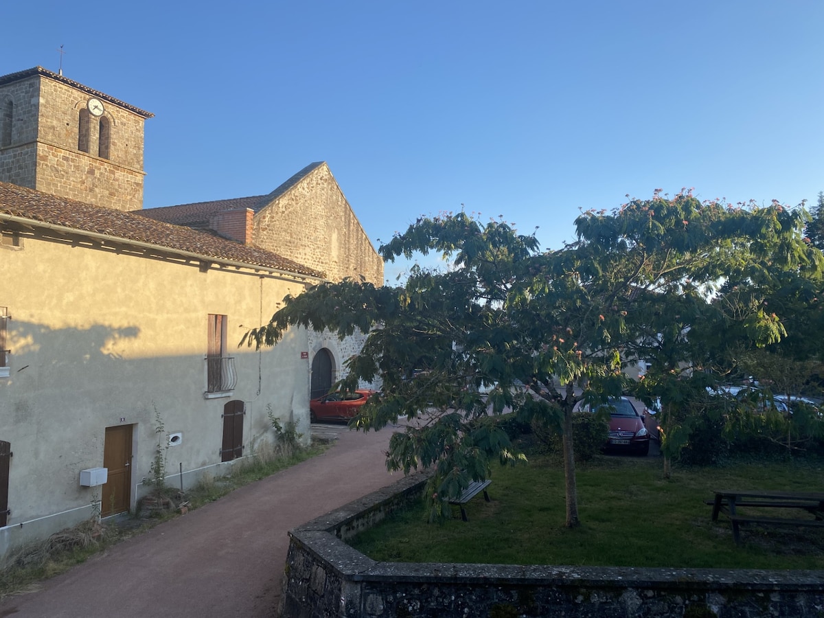 A scene depicting a serene village setting with sunlit buildings and clear skies. A large tree is seen in the foreground, providing greenery to the view. The architecture features a stone church with a clock tower, enhancing the rural charm of the area.