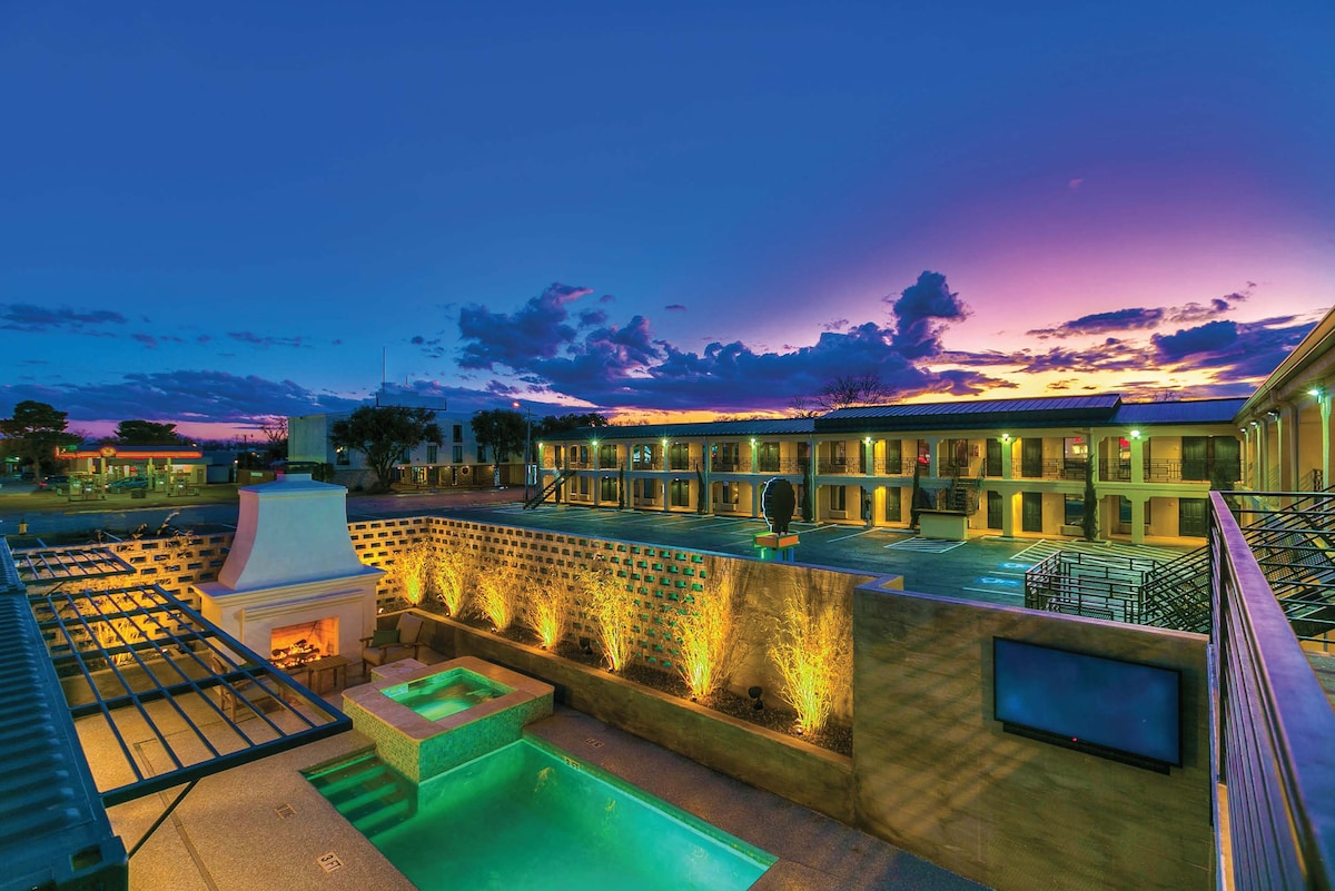 An outdoor area features a serene pool area illuminated by soft lighting. A large, inviting fireplace sits nearby, and the backdrop includes multiple structures with balcony access under a vibrant sunset sky. The scene is complemented by scattered potted plants.