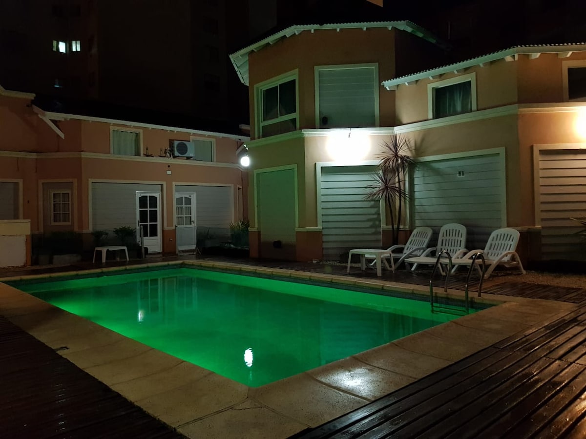 The image showcases a well-lit outdoor pool area at night, featuring a rectangular swimming pool with illuminated turquoise water. Lounge chairs are arranged beside the pool, and the surrounding buildings display large windows and exterior lighting, creating a calm and inviting atmosphere.