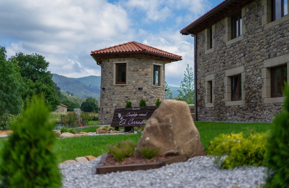 A charming stone building is featured alongside a well-maintained garden area. A prominent sign for 'El Cernado' is displayed in front of the structure. Lush greenery and decorative rocks are arranged strategically on the property, with mountains visible in the background under a partially cloudy sky.