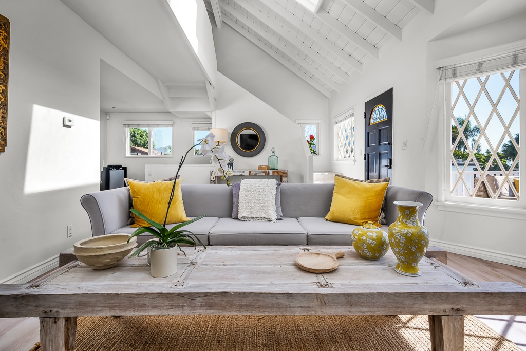 A well-lit living area is centered around a soft gray sofa adorned with two yellow throw pillows. A rustic wooden coffee table, featuring a round tray and decorative items, complements the space. Natural light filters in through large windows, illuminating the vaulted ceiling.