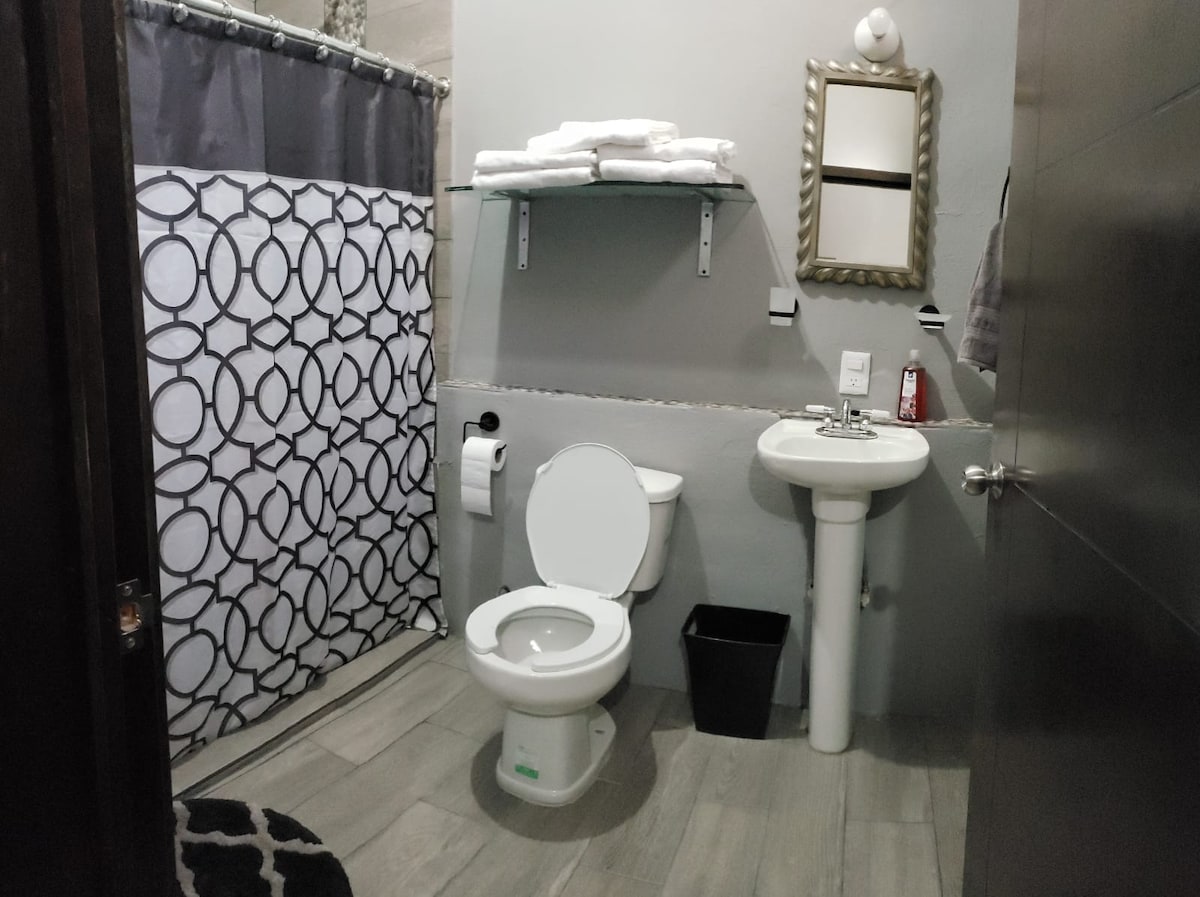 The bathroom features a modern layout with a large shower curtain featuring circular patterns, a white toilet, and a sink with a wall-mounted mirror above. Clean towels are neatly arranged on a shelf, and a small wastebasket is positioned nearby.