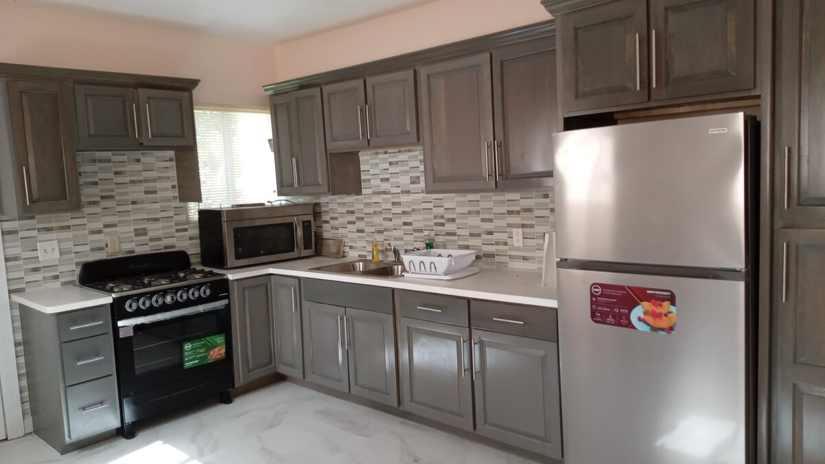 A modern kitchen is equipped with gray cabinetry and a stainless steel refrigerator. A gas stove and microwave are positioned next to a white countertop, which is complemented by a tiled backsplash. Natural light enters through a window near the sink area.