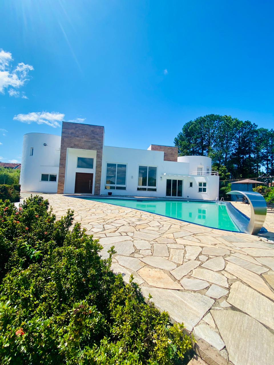 A spacious house is depicted with a heart-shaped pool in front, surrounded by neatly trimmed greenery. The exterior features a combination of white walls and stone accents, while large windows allow natural light to fill the interior. The clear blue sky complements the inviting setting.