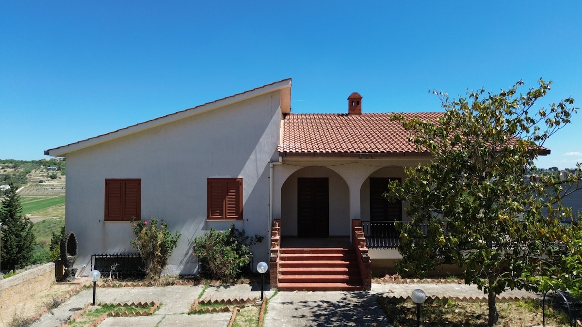 The villa features a spacious exterior with a terracotta-tiled roof and three front steps leading to a covered entrance. Flanking the entrance are two wooden doors beneath a small overhang. Lush greenery and outdoor lighting enhance the property's inviting atmosphere against a clear blue sky.