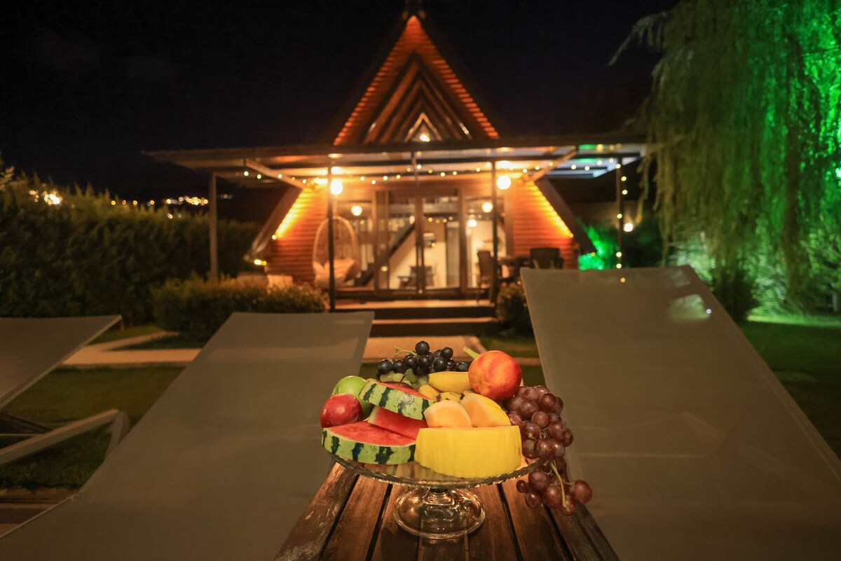 A glass platter filled with a variety of fresh fruits sits on a wooden table, featuring colorful selections like watermelon, grapes, apples, and pineapple. In the background, a well-lit bungalow reflects a welcoming glow, surrounded by greenery and outdoor seating.