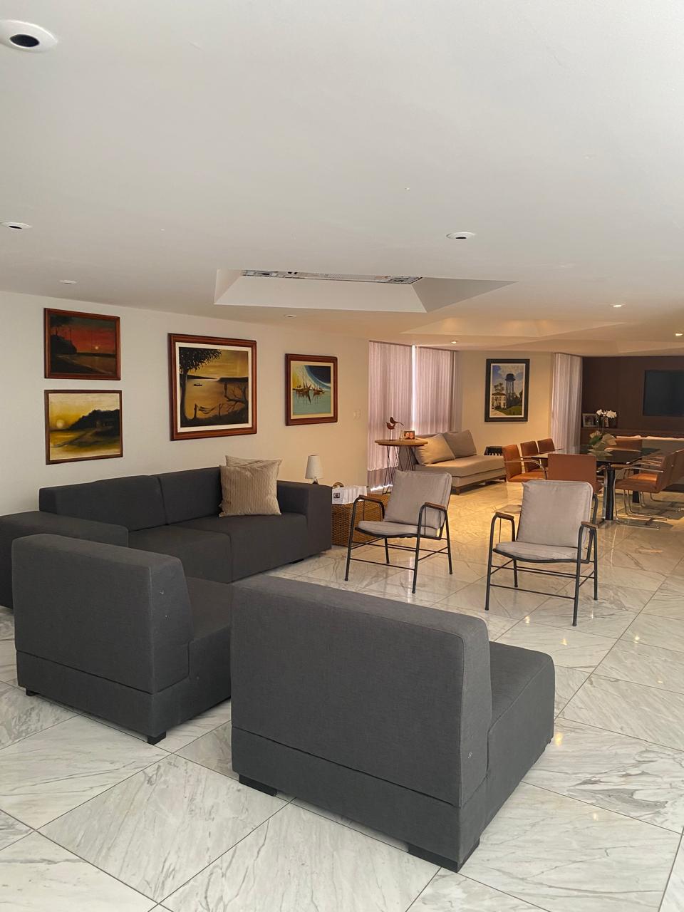 A spacious living area is shown, featuring modern gray sofas arranged around a set of accent chairs. Artwork adorns the walls, and large windows allow natural light to fill the space. The room offers a relaxed atmosphere with a minimalistic design.