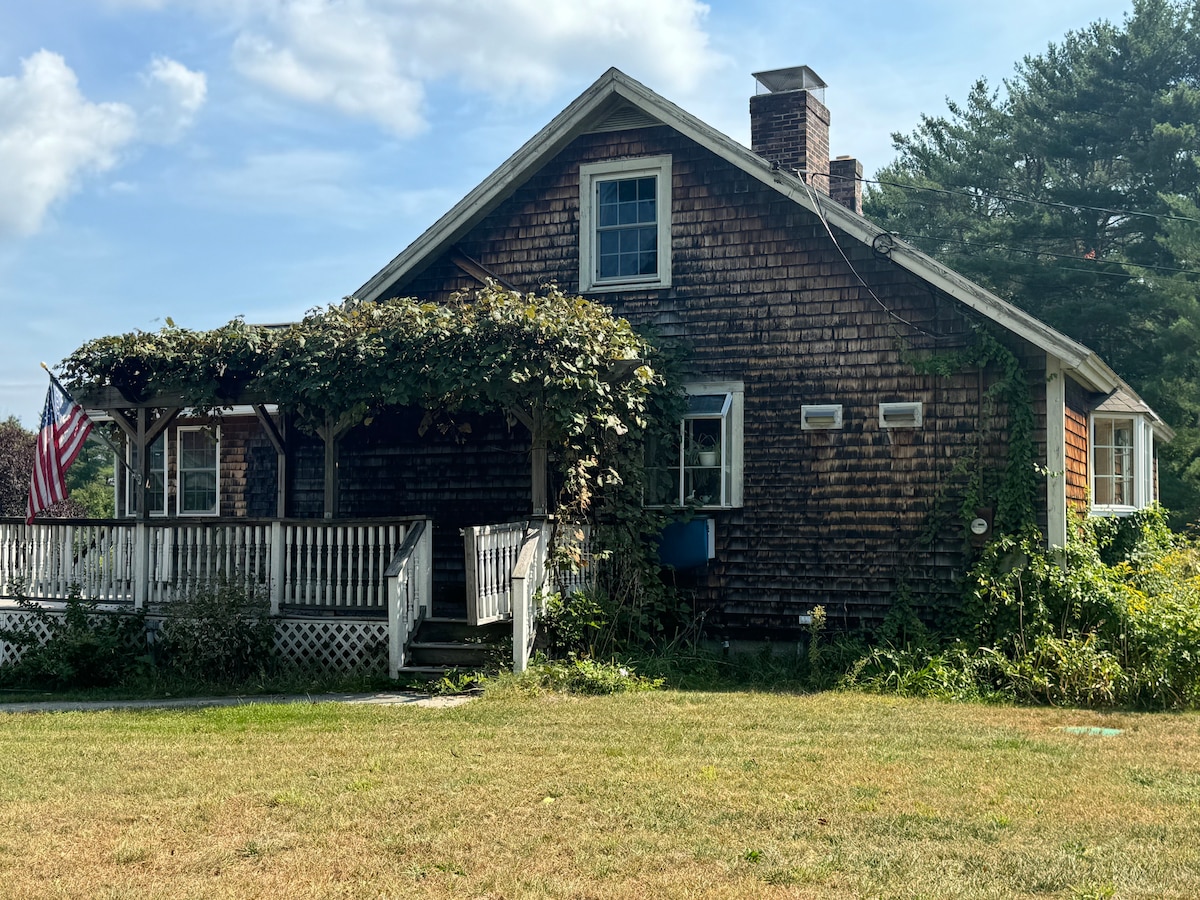 A rustic farmhouse is framed by lush greenery, featuring a welcoming porch adorned with climbing vines. An American flag gently waves, while the wooden exterior showcases a blend of dark and light tones. The yard offers a spacious area, inviting relaxation and outdoor activities.
