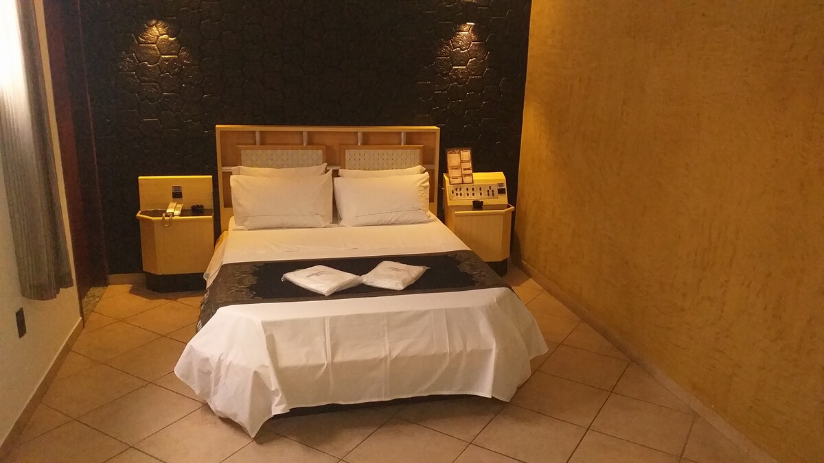 A spacious sleeping area features a large bed with neatly arranged white linens and decorative pillows. Bedside tables with lamps are positioned on either side. The textured walls provide a warm backdrop, and soft lighting enhances the tranquil ambiance.