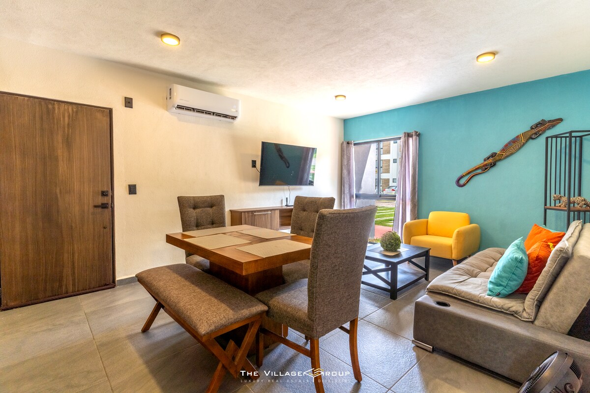 A welcoming living area is presented with a modern dining table surrounded by upholstered chairs. A comfortable sofa with colorful accent pillows is positioned nearby. A large window allows for natural light, while a flat-screen television is mounted on the wall.