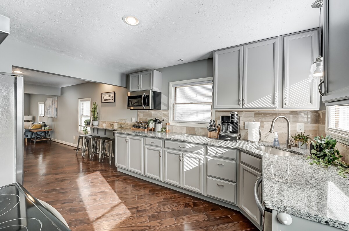 A modern kitchen features light grey cabinetry and granite countertops. Stainless steel appliances are positioned for convenience. A coffee maker and additional kitchenware are visible on the counter. Natural light streams in through multiple windows, illuminating the spacious area.