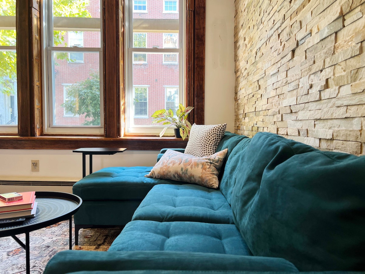 A cozy seating area features a soft teal sectional couch complemented by a decorative pillow. The stone accent wall adds texture, while large windows allow natural light to brighten the space. A coffee table sits nearby, enhancing the inviting atmosphere.