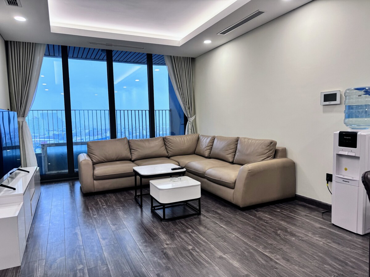 A spacious living area features a large sectional sofa arranged around a modern coffee table. Floor-to-ceiling windows invite natural light and offer a view of the city skyline. A water dispenser and television are positioned nearby, enhancing comfort and convenience.
