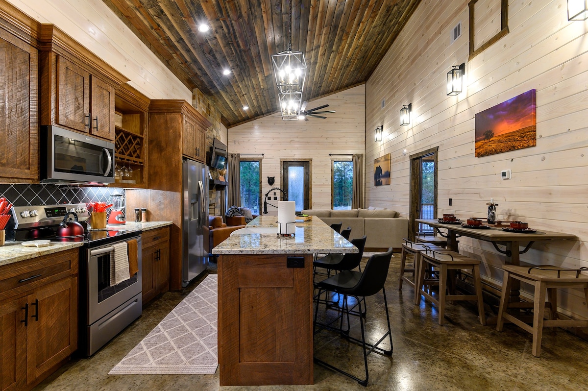 A modern kitchen area is presented with wooden cabinetry and granite countertops featuring bar seating for four. Stainless steel appliances are arranged neatly, while soft lighting illuminates the space. A welcoming dining area with a rustic table and chairs is visible, enhancing the cabin's cozy atmosphere.