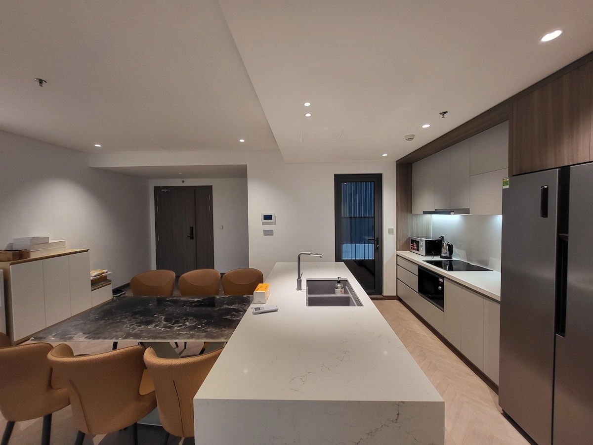 A modern kitchen and dining area feature sleek cabinetry and a large island with a sink. The dining table is surrounded by six beige chairs, and appliances include a refrigerator and a microwave. Natural light enters through a sliding door, which leads to an outdoor space.