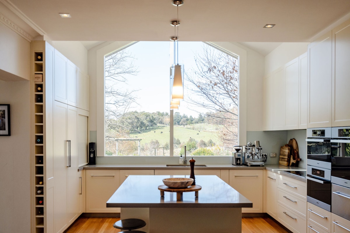 A well-equipped kitchen features modern cabinetry and a central island with seating. Large windows provide natural light and showcase scenic views of the surrounding greenery. Stainless steel appliances are integrated into the design, enhancing the functional and stylish atmosphere.