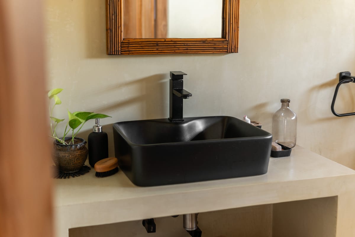 A sleek black basin is positioned on a smooth, neutral-toned countertop. A wooden-framed mirror reflects the space above. Simple bathroom accessories, including a small plant and various bottles, are arranged thoughtfully nearby, contributing to an organized and modern appearance.