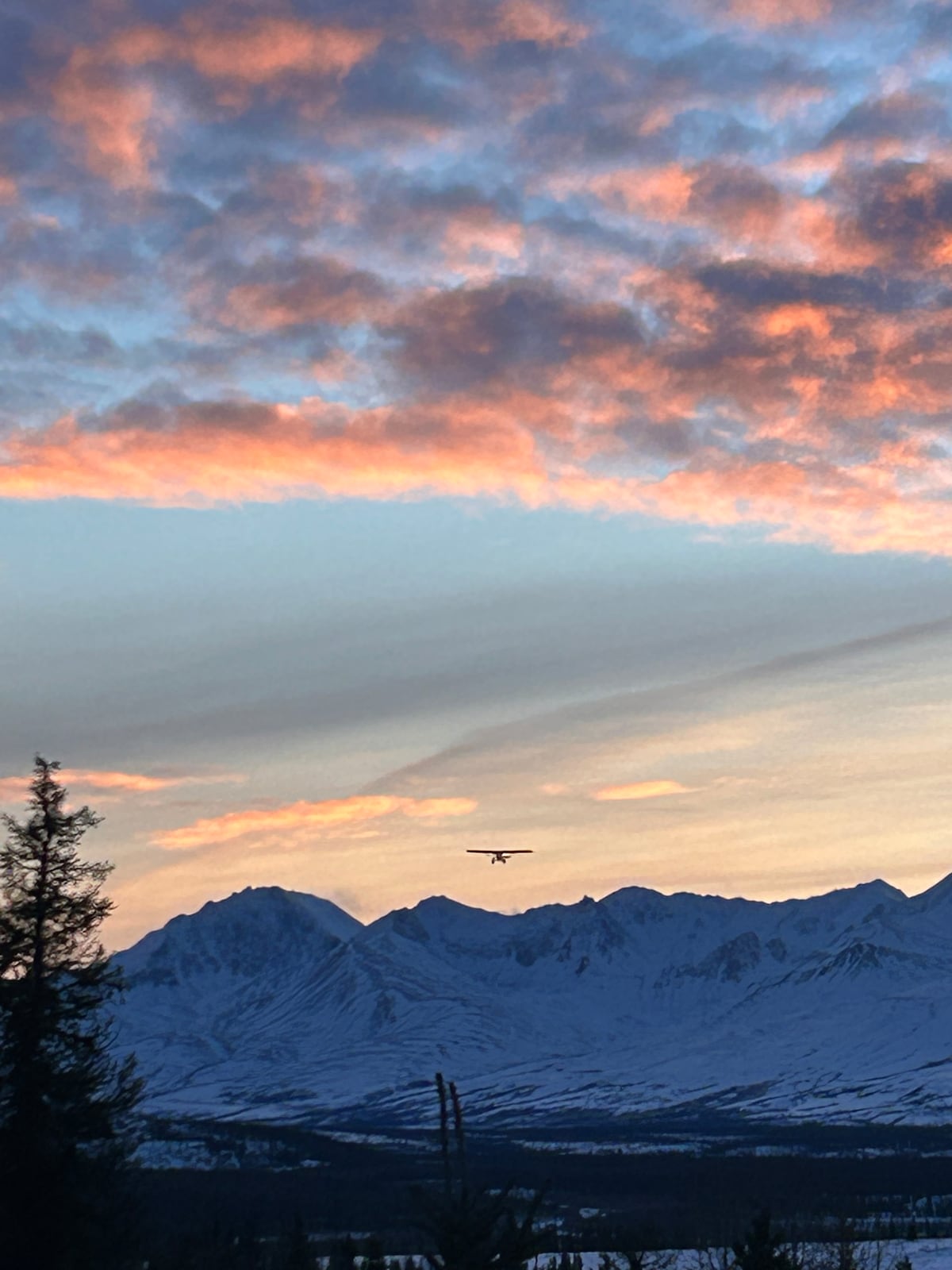 A serene mountain landscape is featured with snow-capped peaks silhouetted against a colorful sunset sky. Soft hues of orange, pink, and purple are blended among gentle clouds. A small airplane is seen flying above the mountains, contributing to the tranquil scene.
