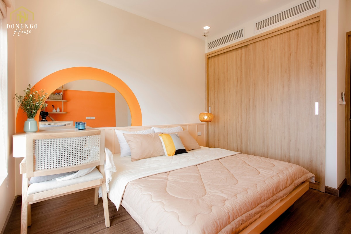 A spacious bedroom features a bed with a light-coloured quilt and several pillows. A curved orange accent wall complements the natural wood accents. A small wooden table with a chair and a decorative plant can be seen in the corner, and a wardrobe is built into the wall.