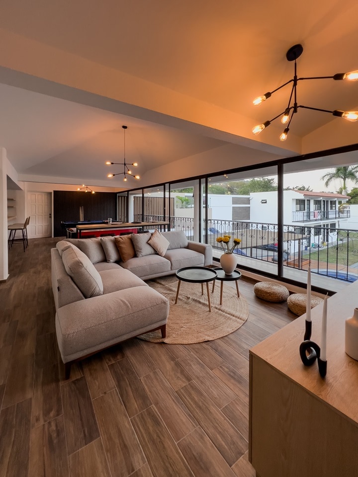 A spacious living area features a soft L-shaped sofa with decorative cushions. A round coffee table sits on a woven rug, while large windows provide natural light and views of the outdoors. A glass dining table is visible in the background, with modern light fixtures overhead.