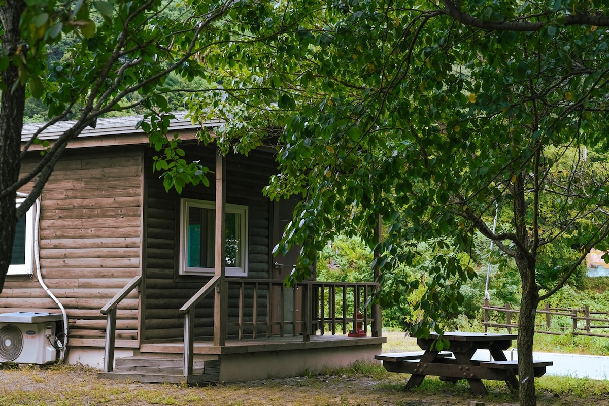A wooden cabin is nestled among trees, featuring a small porch with steps leading to the entrance. A picnic table is situated nearby on the grassy area, surrounded by a peaceful natural environment.