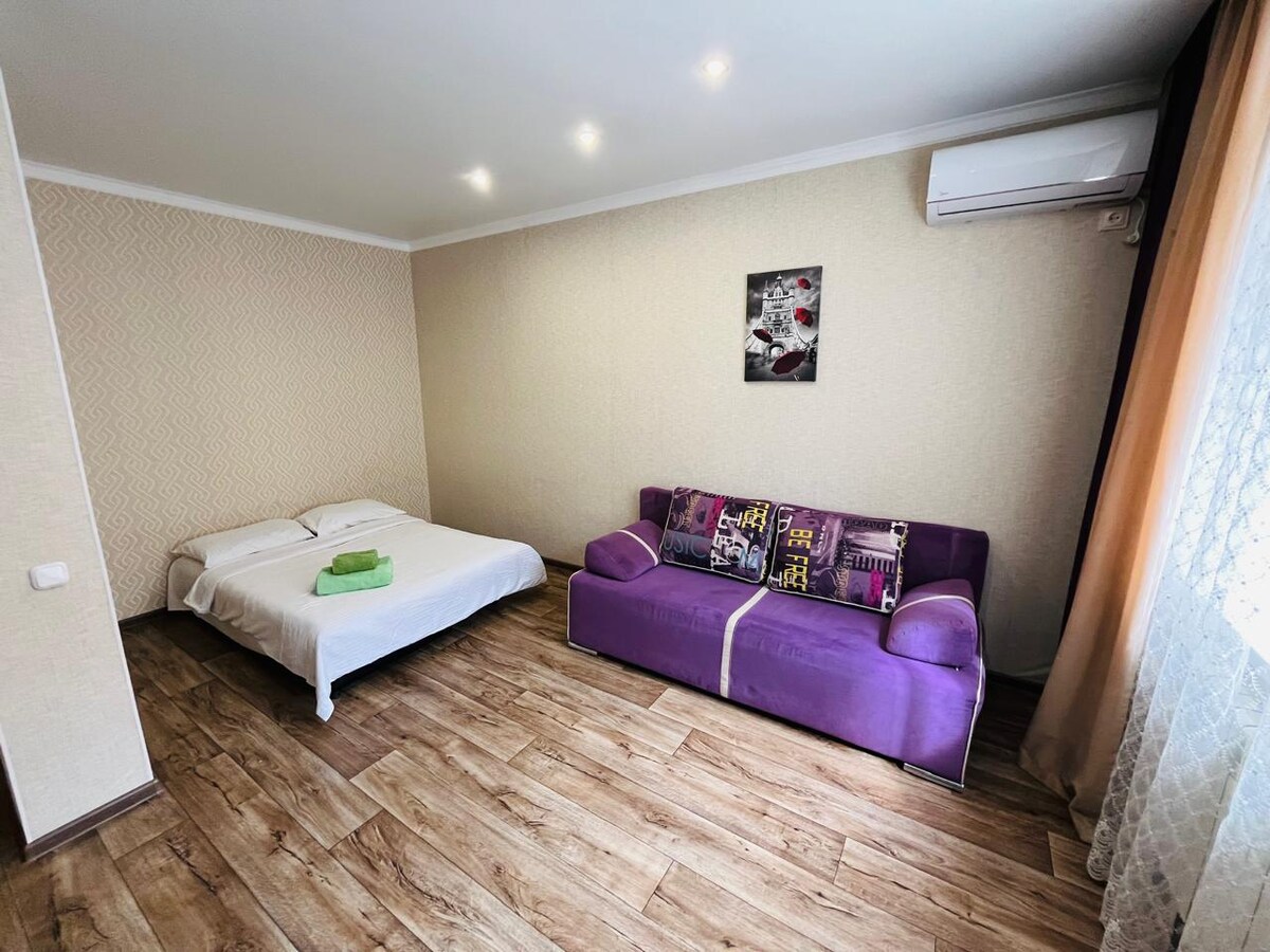 The room features a spacious bed made up with white linens, alongside a purple sofa adorned with decorative pillows. Natural light filters through the sheer curtains, illuminating the wooden floor and a wall with subtle patterned wallpaper.