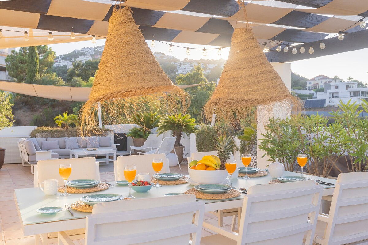 An outdoor dining area is set with a large table covered in plates and glasses of orange drink. Hanging pendant lights and decorative straw elements provide shading. Greenery is visible around the space, complementing a cozy lounge area furnished with cushioned seating.