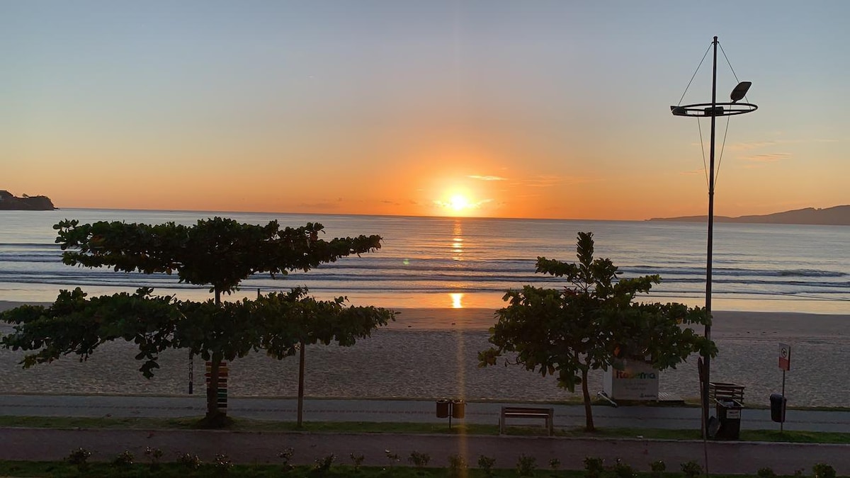 A serene view of the beach at sunrise, featuring gentle waves lapping at the shore. Silhouetted trees frame the scene, with a nearby lamp post standing beside the beach walkway. The soft colors of the sky reflect on the water, creating a tranquil atmosphere.