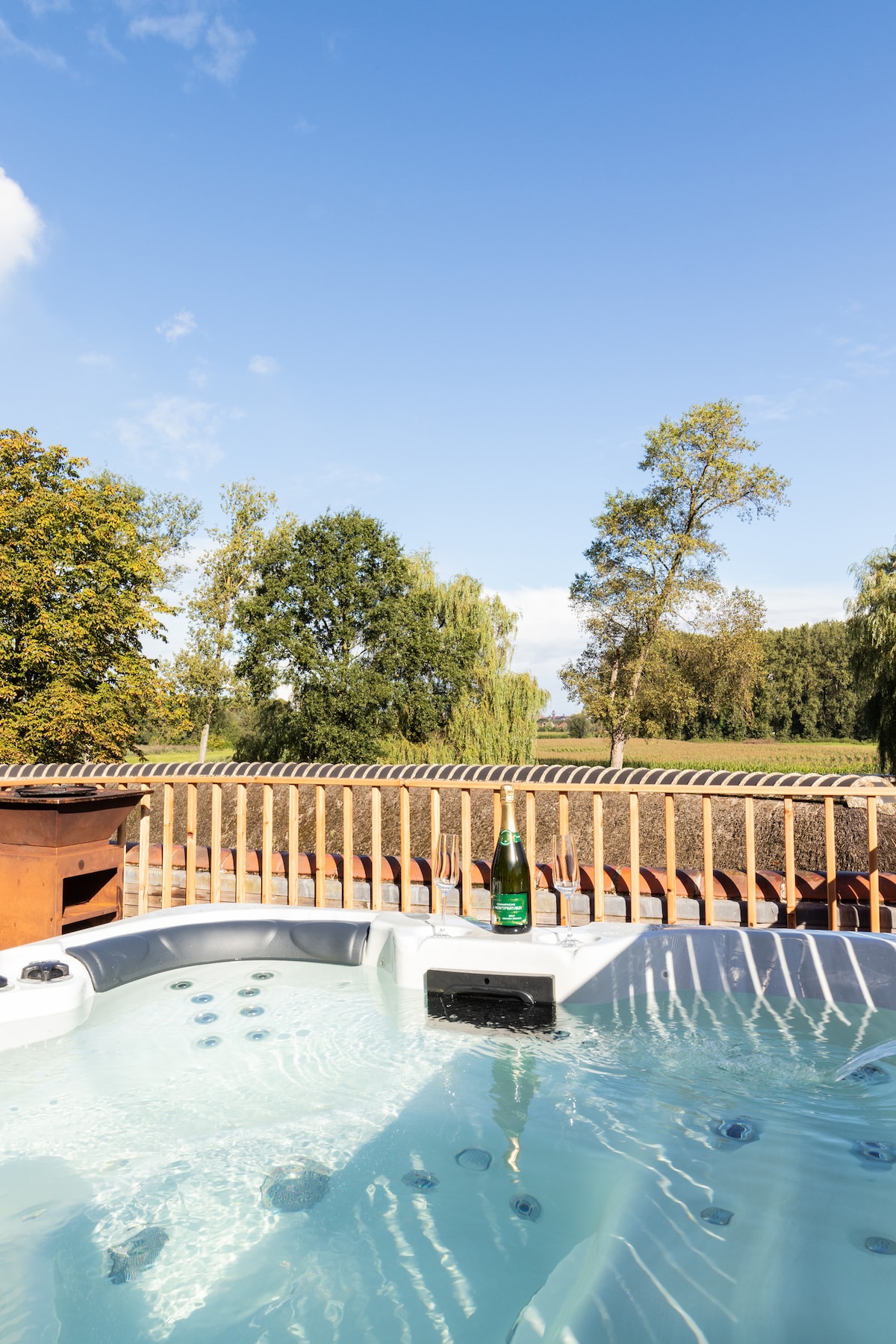A private jacuzzi is positioned on a terrace with wooden railings, overlooking lush greenery and trees. A bottle sits on the edge of the jacuzzi, reflecting a clear blue sky. The tranquil setting invites relaxation and enjoyment of the natural surroundings.