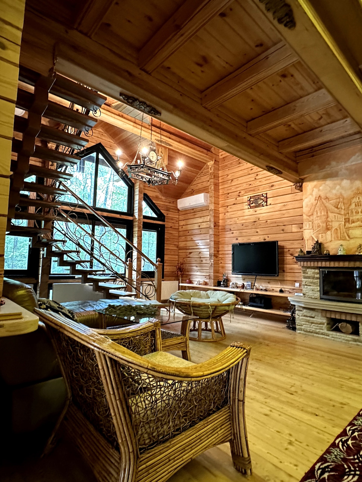 An interior space features wooden walls and a staircase with an ornate railing. A cozy seating area includes woven chairs and a television mounted above a stone fireplace. Natural light enters through large windows, accentuating the warm tones of the wooden decor.