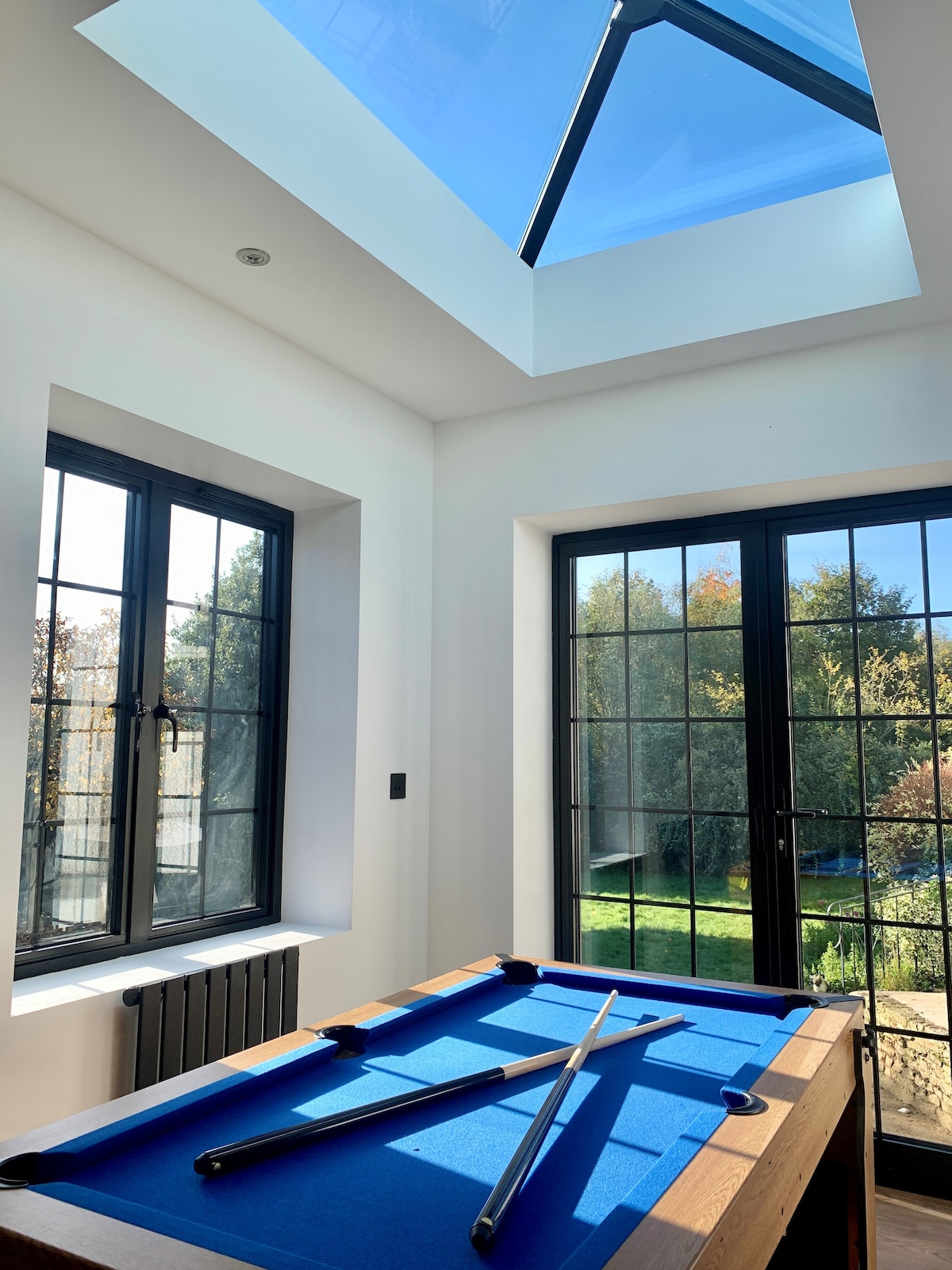 A bright game room features a blue billiard table positioned beneath a skylight. Large windows framed in black provide views of the lush greenery outside, allowing natural light to flood the space.