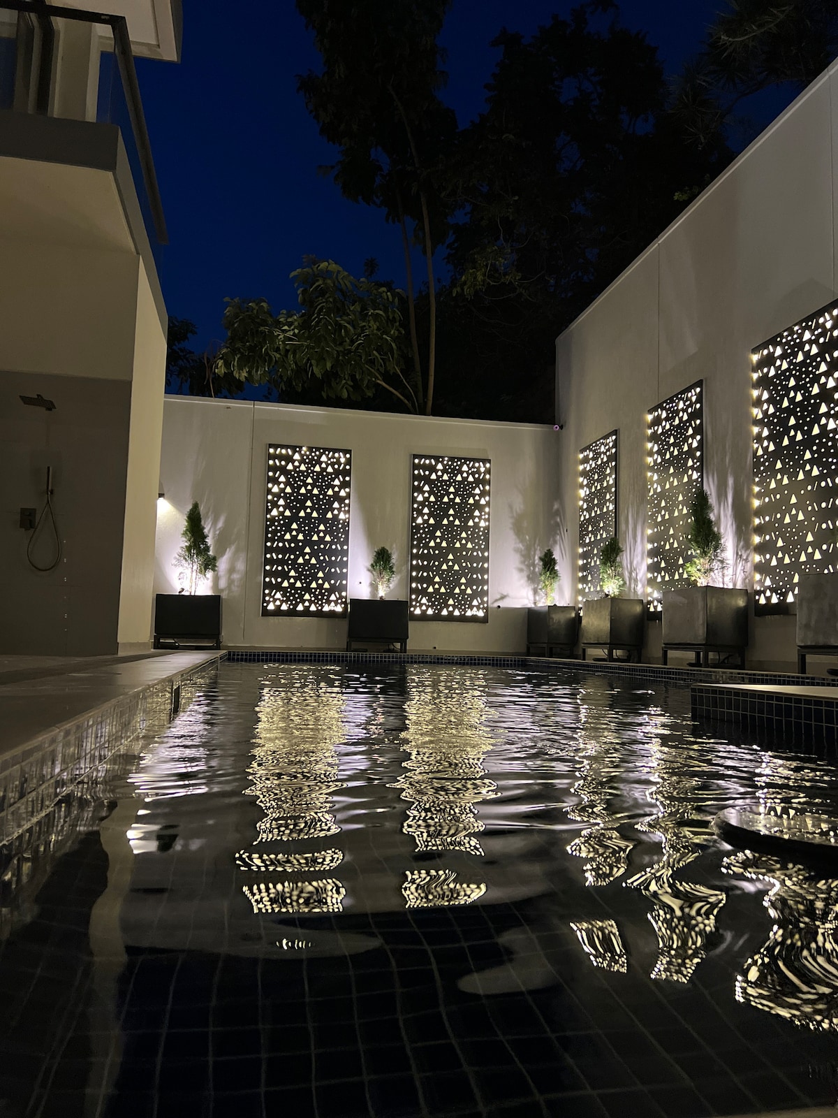 An illuminated pool reflects soft light from decorative screens, creating a serene nighttime atmosphere. Lush greenery can be seen in the background, while minimalist outdoor seating adds to the inviting setting.