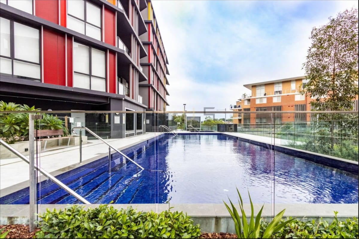 A modern swimming pool is framed by contemporary buildings and lush greenery. The clear water reflects the sky, while stylish glass fencing enhances the open feel. Paved areas and neatly arranged plants surround the pool, creating a welcoming outdoor environment.