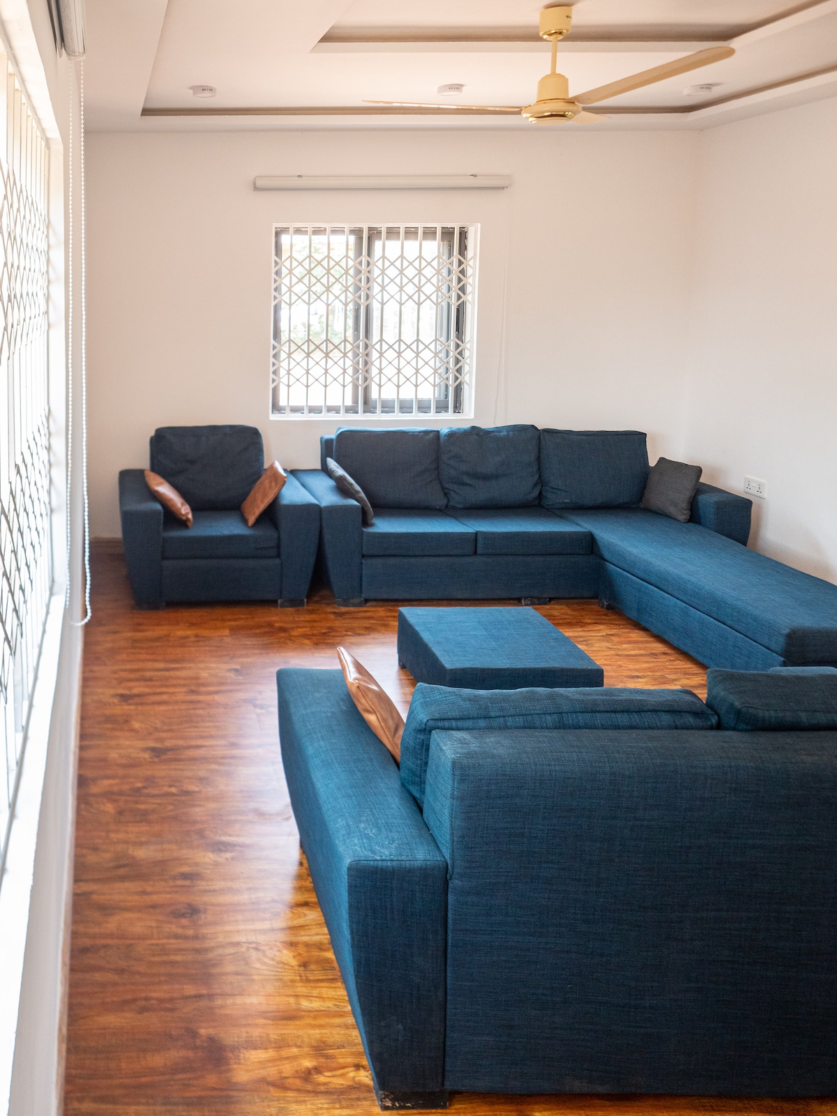 A spacious living area is furnished with a blue sectional sofa, complemented by accent chairs. A central coffee table unifies the seating arrangement. Natural light filters through the window, highlighting the warm wooden floor and the simplicity of the space.