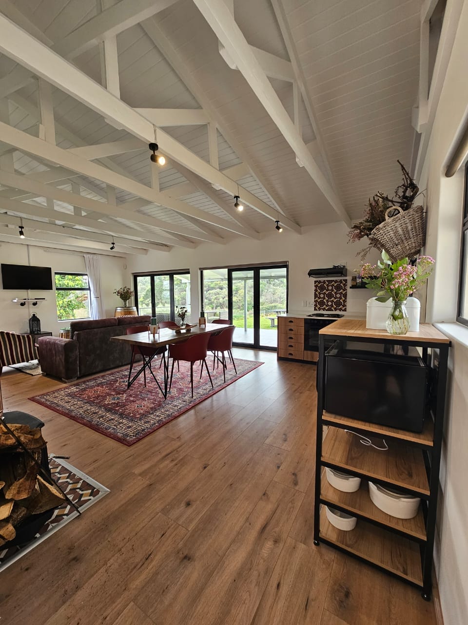An open living space is highlighted by high white-beamed ceilings and large glass doors, providing access to outdoor views. A cozy seating area is complemented by a modern dining table for four. A decorative rug adds warmth near the fireplace, with wood storage beside it.